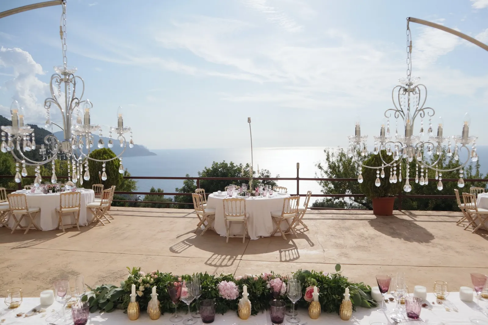 Mesa de boda vestida de blanco en una terraza sobre el mar, con dos lámparas de araña de cristal colgadas de báculos curvos