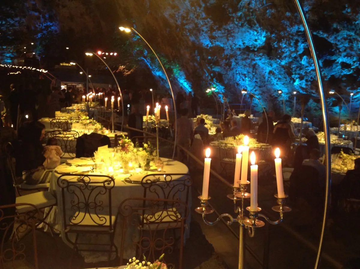 Mesas redondas iluminadas con candelabros en una cena nocturna al aire libre, entre árboles iluminados en azul