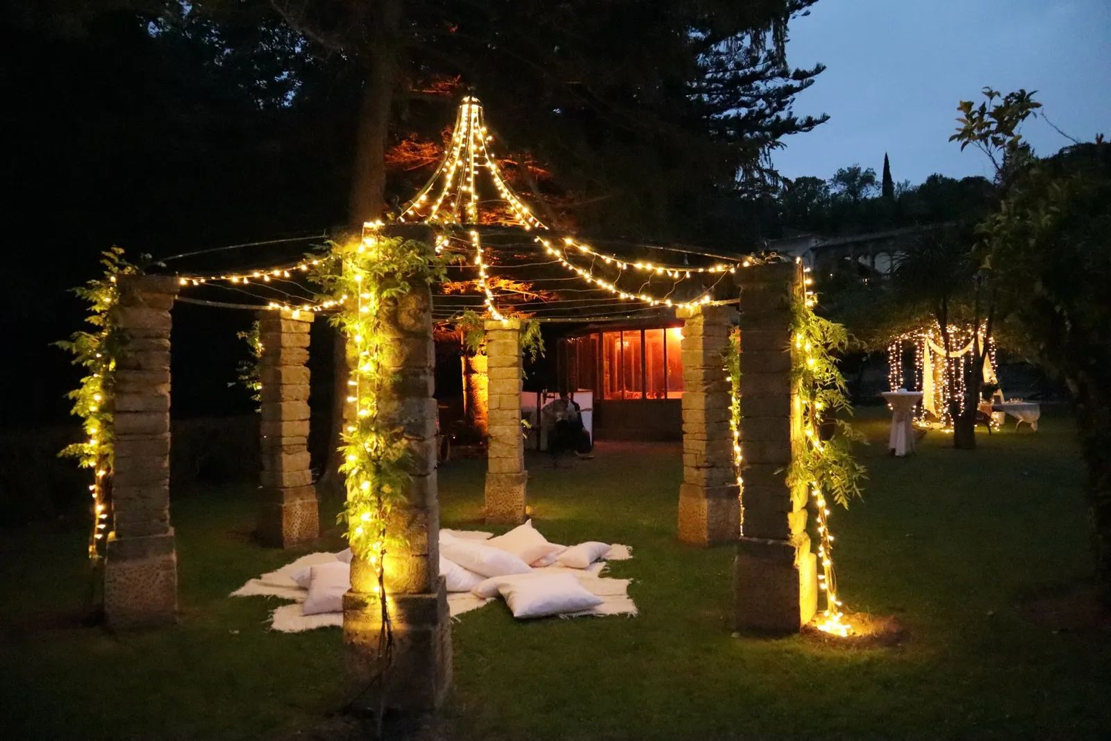 Pérgola de piedra decorada con guirnaldas de bombillas y vegetación, con cojines sobre el césped