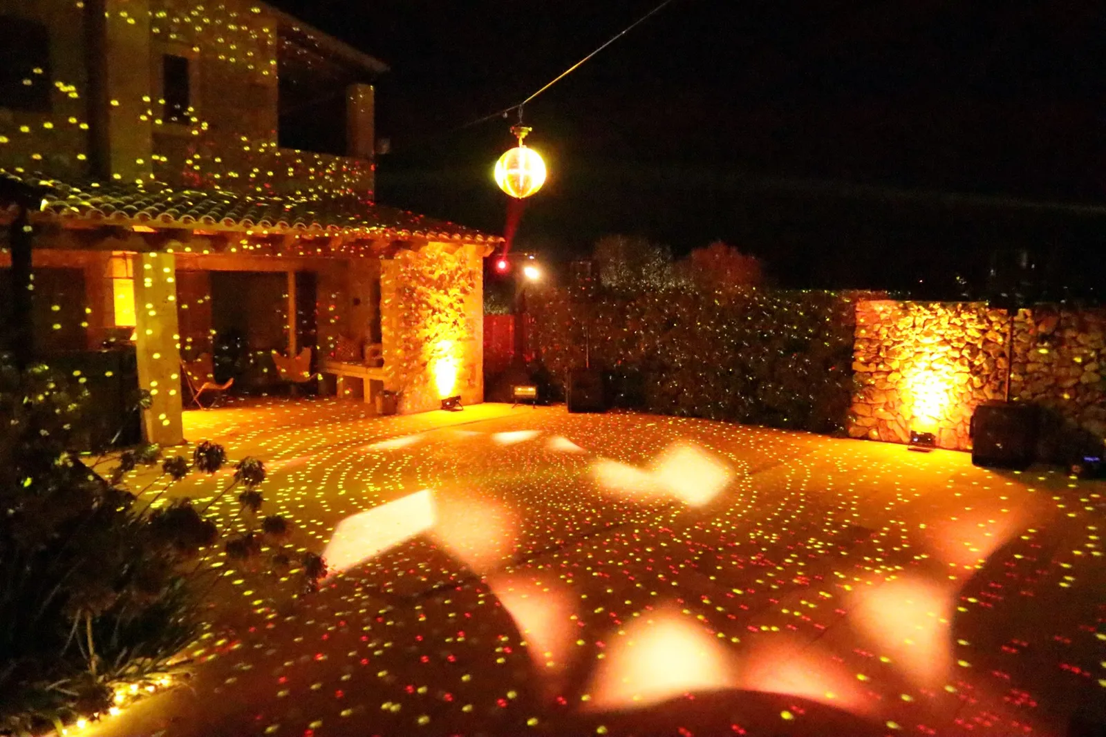 Patio de una casa de piedra con guirnaldas en la fachada, una bola de espejos y puntos de luz proyectados sobre el suelo