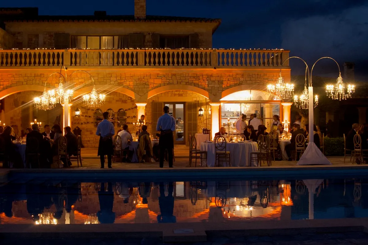 Evento nocturno junto a una piscina, con los arcos de una casa señorial bañados en luz ámbar y arañas colgadas sobre las mesas